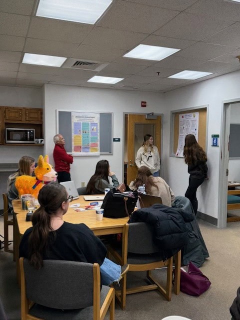 A classroom scene where students are seated at tables while two individuals present posters on the wall. The posters include colorful educational content related to mathematics. A bright orange plush animal is visible on a table, along with notebooks and drinks.

