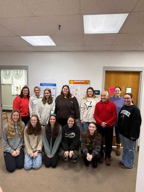 A group photo of students standing and kneeling in two rows inside a classroom. Behind them, educational posters are displayed on the wall, including one labeled “Geometry.” The setting includes a door and bulletin board.
