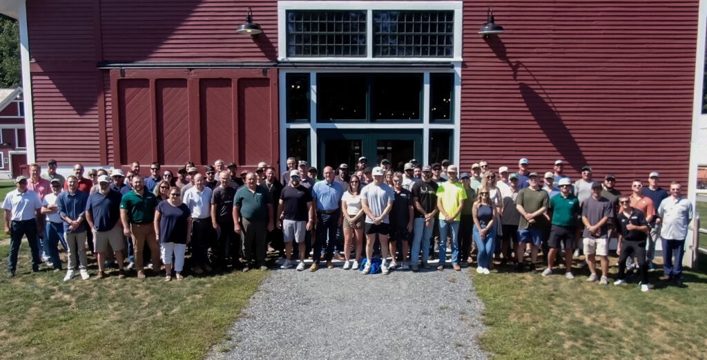 Large group of people standing together outdoors in front of a tall red barn-style building on a sunny day.