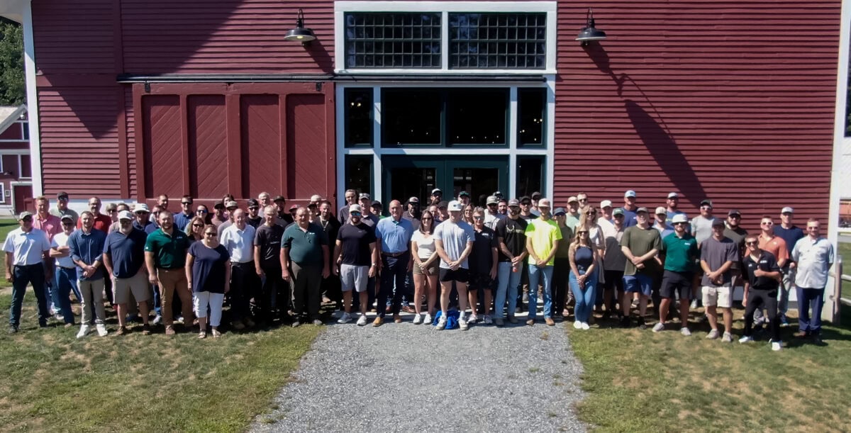 Large group of people standing together outdoors in front of a tall red barn-style building on a sunny day.
