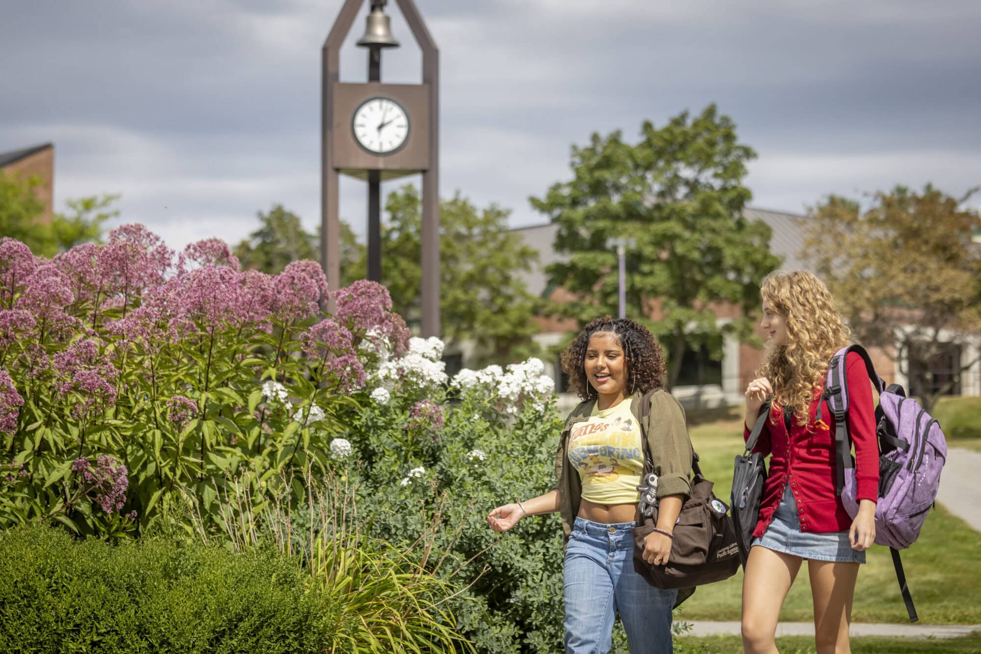 Two young women walking past floral bushed on the VTSU Johnson campus, a large clock tower is in the background