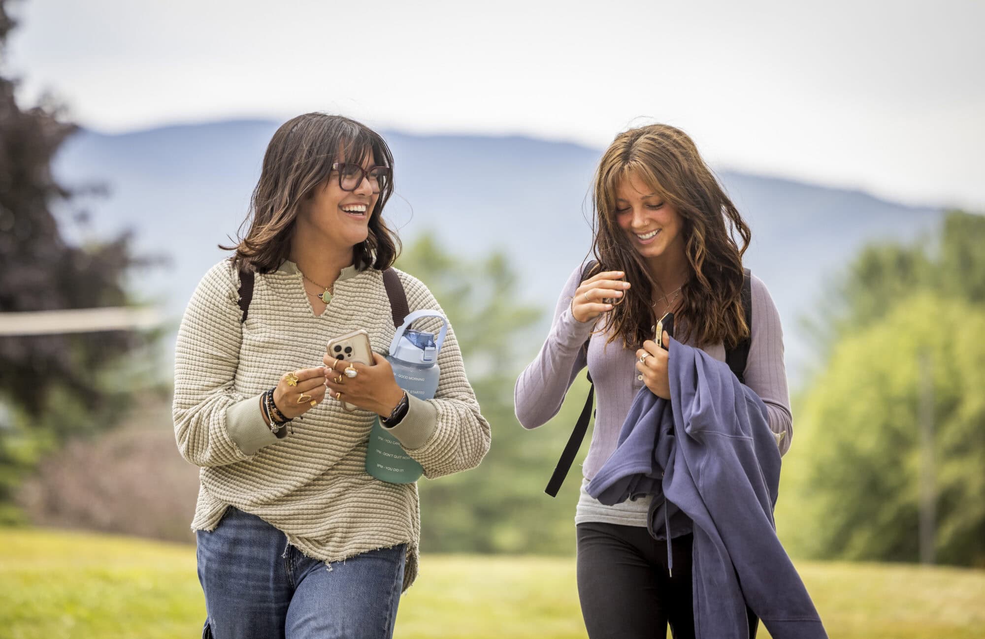 Two young women walking through a grassy area on teh VTSU Johnson campus. Trees and mountains can be seen in the background.
