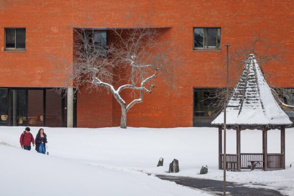 A snowy campus scene with a red brick building in the background. Two people wearing winter coats walk along a snow-covered path on the left. In the center, a leafless tree is dusted with snow. On the right, a small wooden gazebo with a snow-covered roof stands beside a cleared walkway.