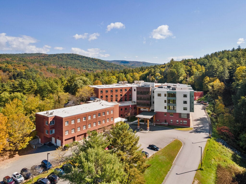 Aerial view of a multi‑story brick and white building surrounded by trees with autumn foliage, set against rolling hills under a blue sky.