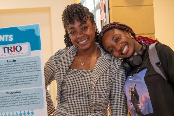 Two young women standing by a research poster and smiling, one is resting her head on the others shoulder