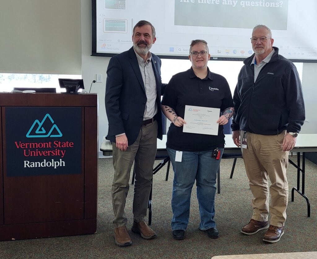 Three people stand in a classroom at Vermont State University Randolph, with the person in the center holding a certificate while standing between two presenters near a podium.
