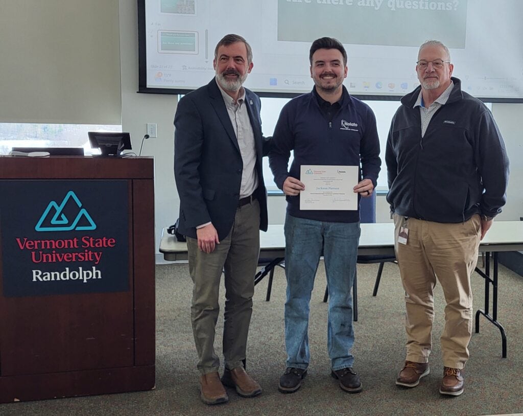 Three people stand in a classroom at Vermont State University Randolph, with the person in the center holding a certificate while standing between two presenters near a podium.
