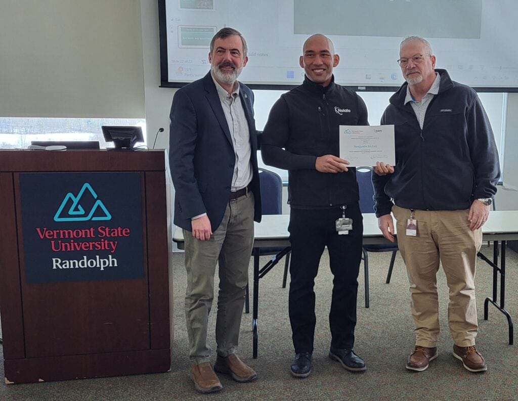 Three people stand in a classroom at Vermont State University Randolph, with the person in the center holding a certificate while standing between two presenters near a podium.
