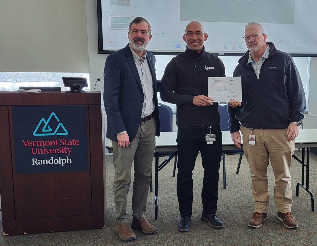 Three people stand in a classroom at Vermont State University Randolph, with the person in the center holding a certificate while standing between two presenters near a podium.