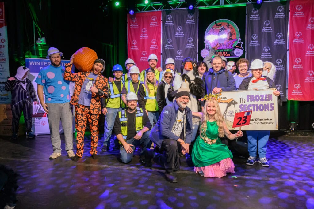 Group of people in colorful costumes and safety vests posing on a stage with bright lights and Special Olympics banners behind them; one person holds a sign labeled ‘The Frozen Sections 23.