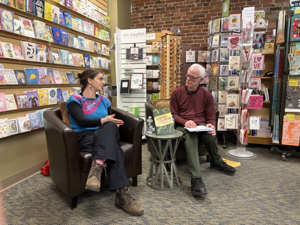 Two people sit in armchairs inside a cozy bookstore, engaged in what appears to be a book discussion or author event. They are seated near a small round table holding a book, a water bottle, and papers. Behind them are shelves filled with colorful greeting cards, books, and art supplies. One person is speaking with hands gesturing, while the other holds an open book on their lap. The space has warm lighting, exposed brick walls, and a welcoming, creative atmosphere.
