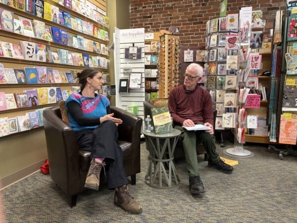 Two people sit in armchairs inside a cozy bookstore, engaged in what appears to be a book discussion or author event. They are seated near a small round table holding a book, a water bottle, and papers. Behind them are shelves filled with colorful greeting cards, books, and art supplies. One person is speaking with hands gesturing, while the other holds an open book on their lap. The space has warm lighting, exposed brick walls, and a welcoming, creative atmosphere.