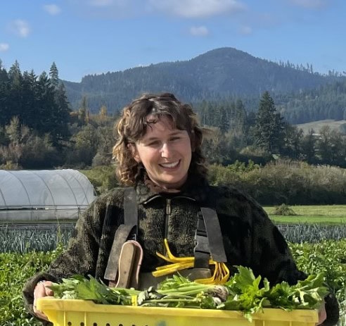 Taylor Paone standing at the farm on the VTSU Randolph campus and holding a basket full of produce while smiling at the camera