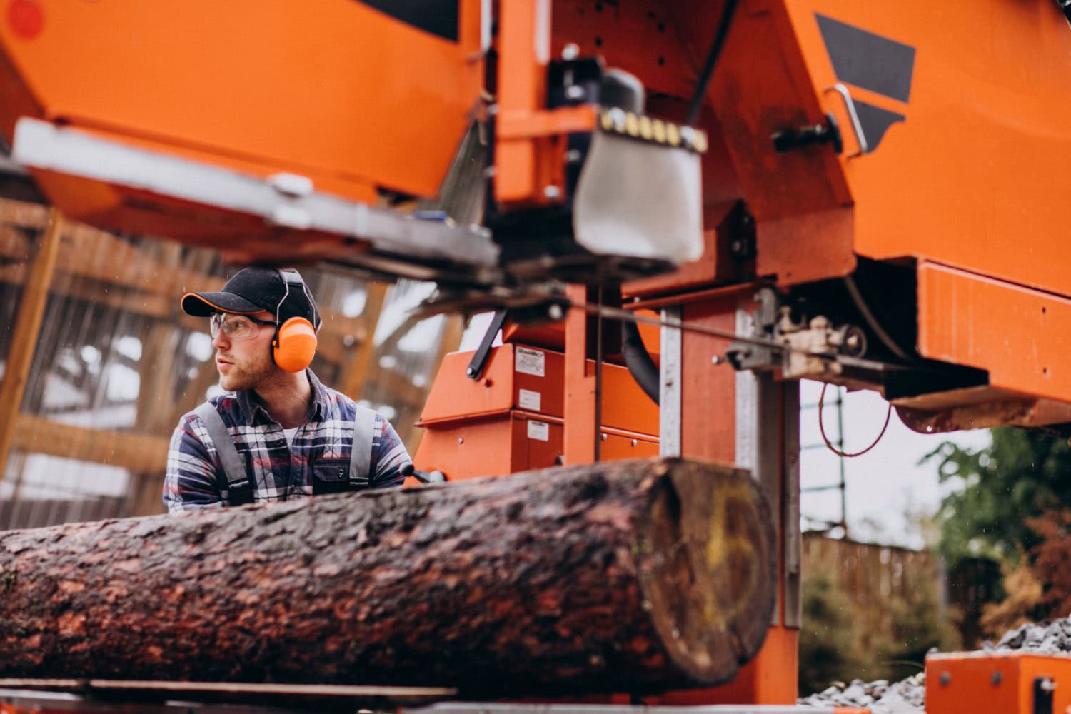 A man wearing a flannel shirt, ball cap, and protective earmuffs pushing a large log down a machine at a sawmill.