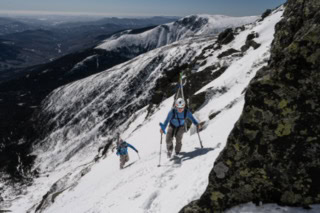 Two people climb up a narrow, snowy alpine ridge carrying skis on their backpacks, with rugged mountain peaks spreading out behind them.