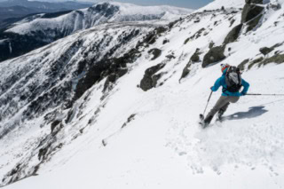 A person in a teal jacket skis down a wide, steep mountainside with expansive snowfields and distant ridgelines stretched across the horizon.