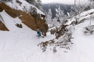 A person in a teal jacket skis down a tight, rocky couloir with snow-covered cliffs on both sides and a forested valley visible below.