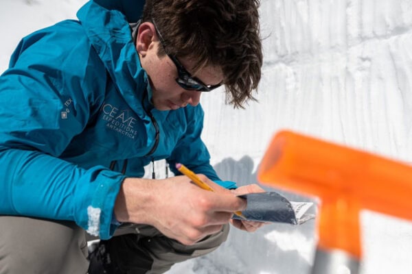 A person in a teal jacket kneels beside a cleanly cut snowpit, taking notes on a small notebook while holding a pencil, with avalanche assessment tools visible nearby.