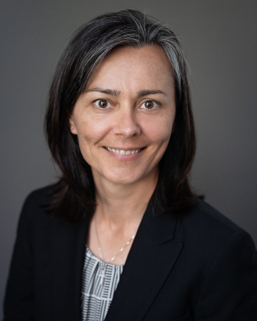 A person wearing a dark blazer over a patterned top is posed against a plain, neutral-colored background. The person has straight, shoulder‑length dark hair with lighter strands near the top.