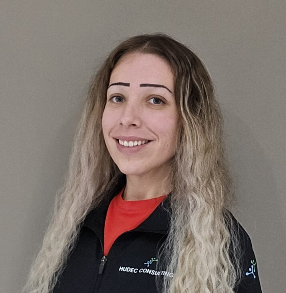 Portrait of a person standing against a plain beige wall, wearing a black zip-up jacket over a red shirt. The jacket has the visible text “HUDEC CONSULTING” on the left chest, and the person has long, wavy, light-colored hair and is facing the camera.