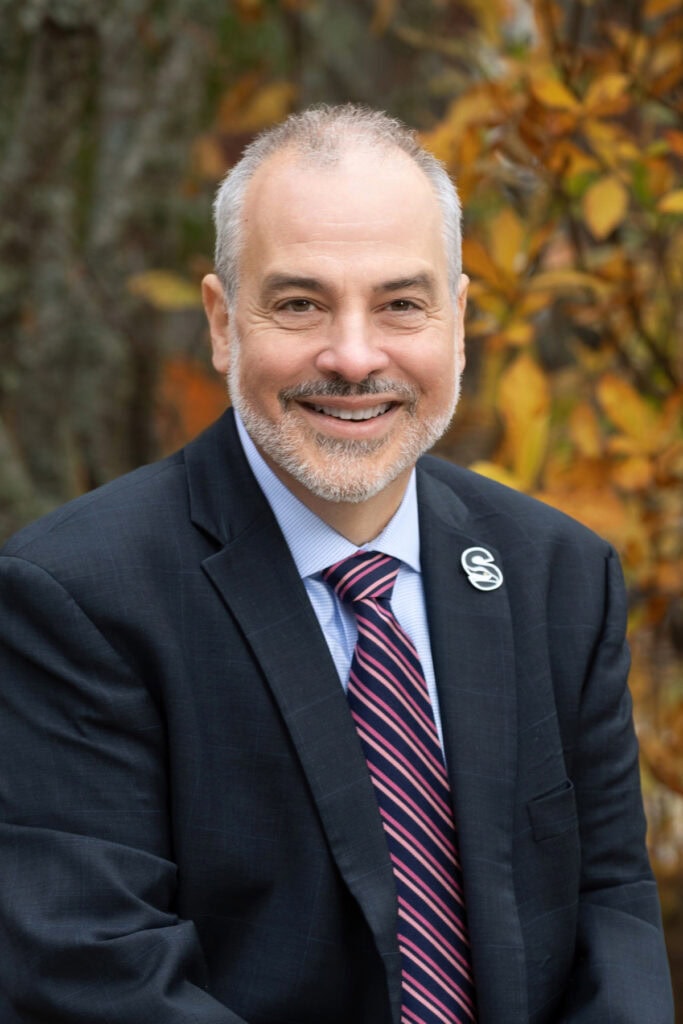 Person in a dark suit jacket, light dress shirt, and striped tie, smiling while seated outdoors with autumn foliage in the background.
