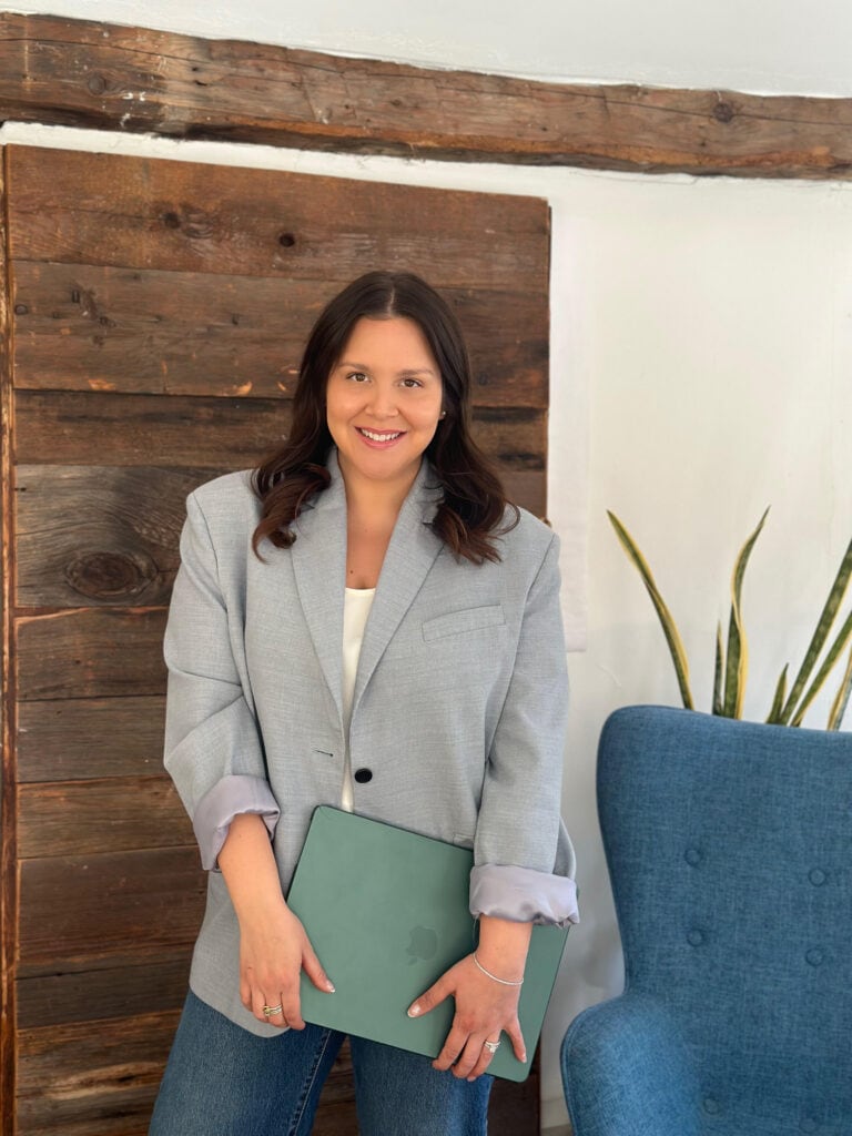 Smiling person standing indoors in front of a rustic wooden wall, wearing a light gray blazer and jeans, holding a closed green laptop, with a blue chair and tall plant nearby.