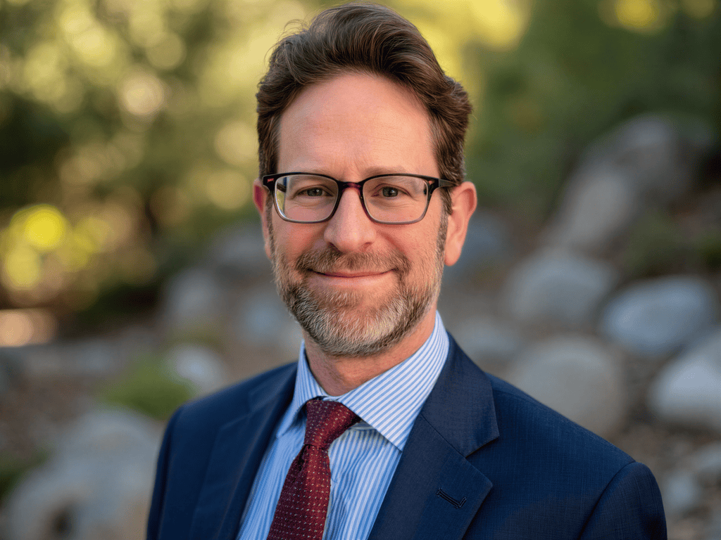 Headshot of a man wearing glasses, a navy suit, light blue striped shirt, and red tie, smiling slightly outdoors with blurred greenery and rocks in the background.