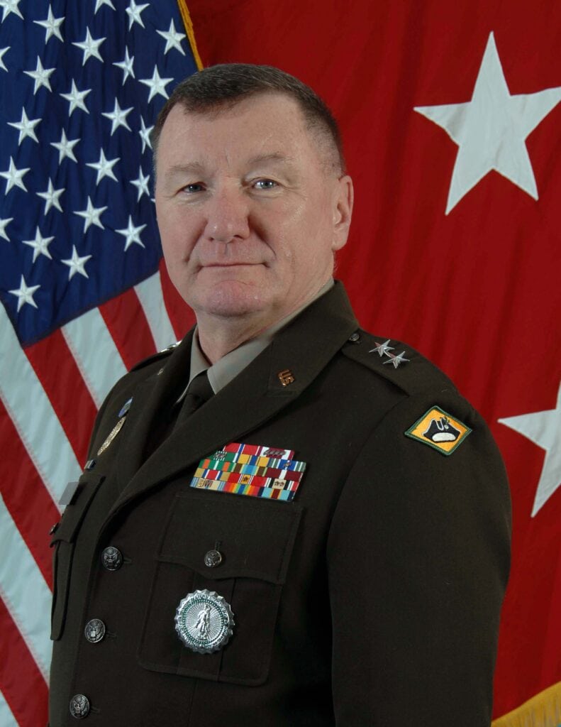 Person in a dark military dress uniform with medals and insignia, posed in front of an American flag and a red flag with a white star.