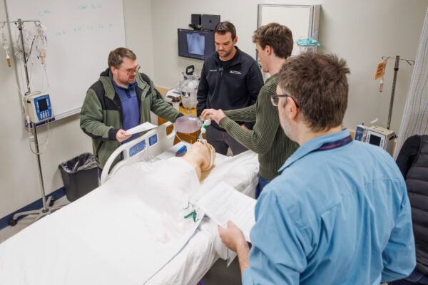 Four adults stand around a hospital bed in a clinical lab setting, reviewing papers and handling medical equipment while practicing a procedure on a patient simulation mannequin. The room contains IV poles, monitors, tubing, and a whiteboard, indicating a hands-on medical or healthcare training scenario.