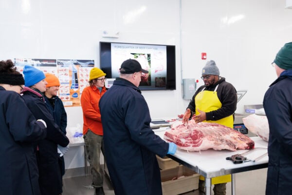 Participants gather around a stainless‑steel table inside VTSU’s 1787 Butchery as an instructor demonstrates meat cutting on a large cut of meat; attendees wear cold‑weather clothing and food‑safe aprons, with instructional posters and a wall‑mounted screen visible in the background.