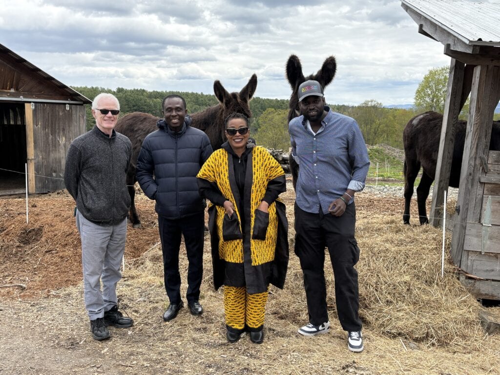 Four people posing outdoors at a farm with two dark-colored donkeys behind them, surrounded by hay and farm structures under a partly cloudy sky.