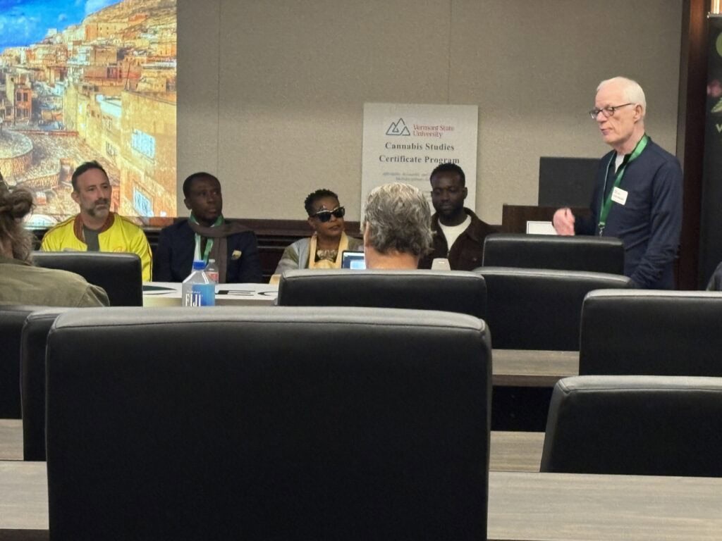 A panel discussion in a conference room with four panelists seated at a table and a moderator standing to the right, with a Vermont State University Cannabis Studies Certificate Program sign visible in the background and a colorful cityscape projected on a screen.