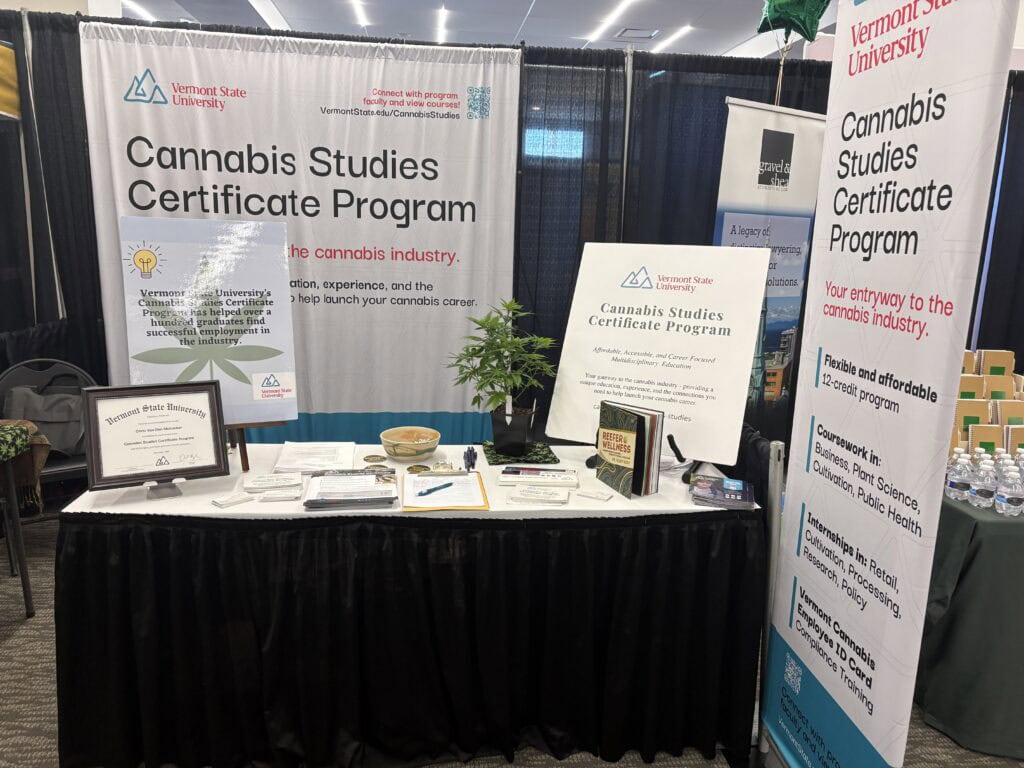 A trade show booth for Vermont State University's Cannabis Studies Certificate Program, featuring banners, a small cannabis plant, informational materials, a framed certificate, and the book Reefer Wellness on display.