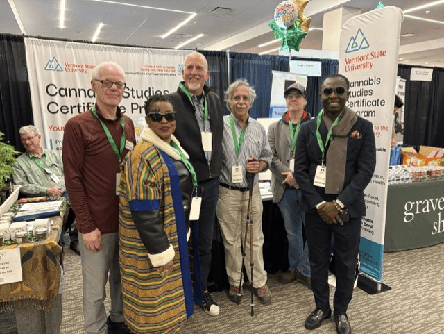 Six people posing together in front of the Vermont State University Cannabis Studies Certificate Program trade show booth at a convention.