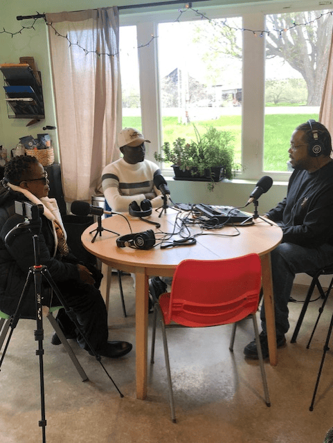 Three people seated around a small round table recording a podcast, with microphones, headphones, and audio equipment visible in a cozy, well-lit room.