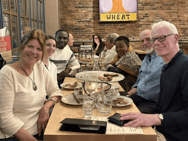 A group of eight people sharing a meal at a long table inside a casual brick-walled restaurant.