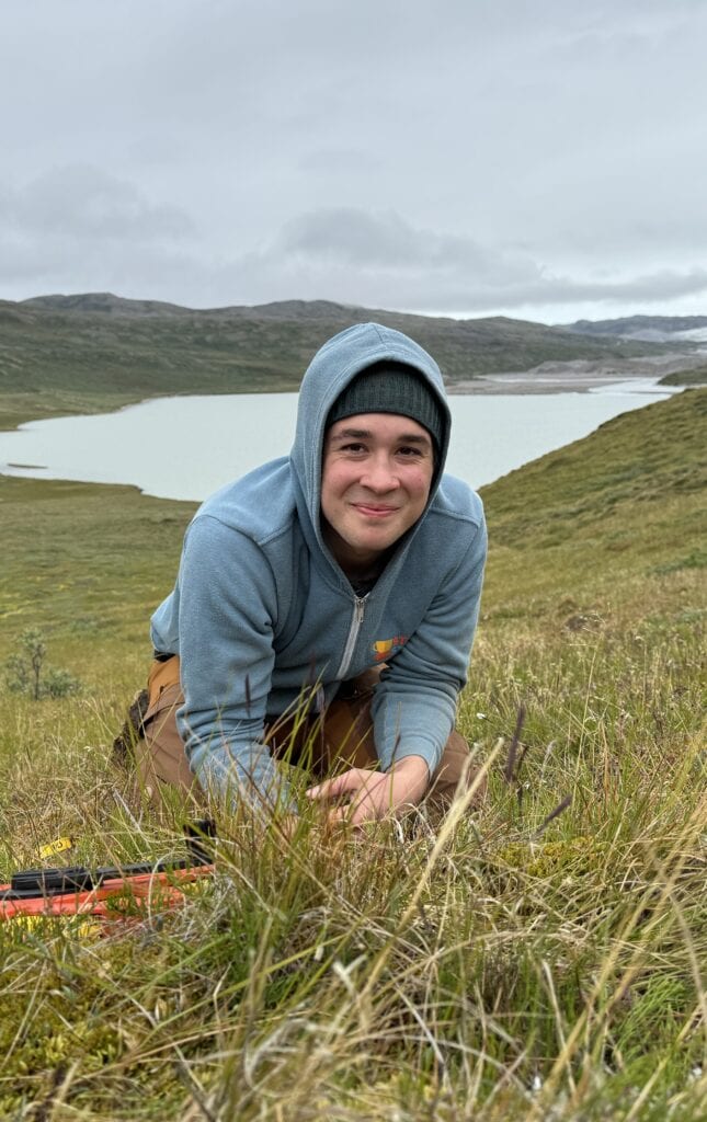 Dominic Mazzilli smiling at the camera wearing a blue hoodie and black hat while crouching on a grassy hill overlooking a lake.