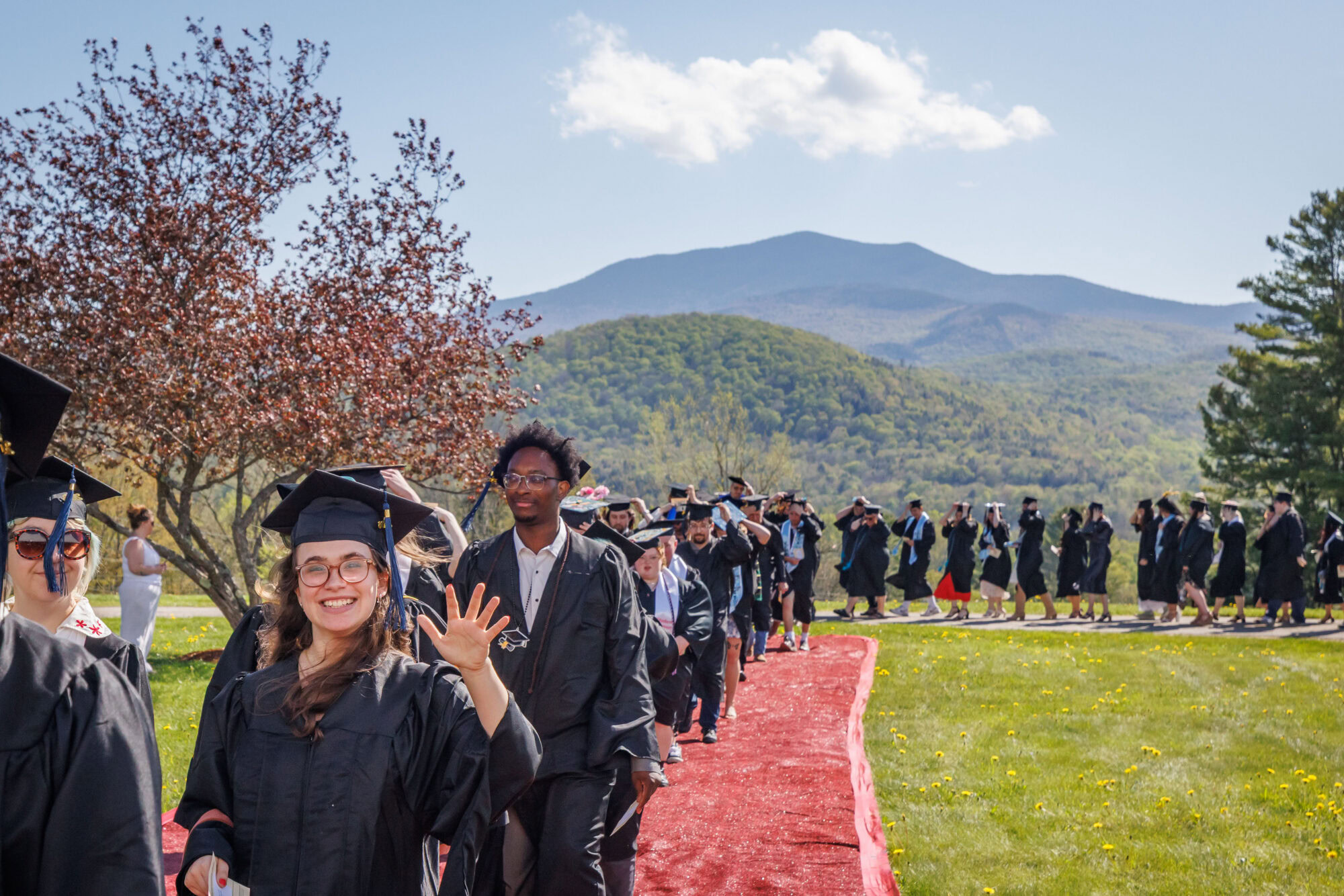 A smiling graduate waves to the crowd while processing along a red carpet pathway during an outdoor commencement ceremony. A long line of fellow graduates in black caps and gowns stretches behind her across a green hillside, with a forested Vermont mountain rising prominently under a blue sky in the background.