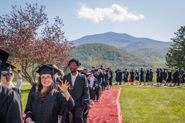 A smiling graduate waves to the crowd while processing along a red carpet pathway during an outdoor commencement ceremony. A long line of fellow graduates in black caps and gowns stretches behind her across a green hillside, with a forested Vermont mountain rising prominently under a blue sky in the background.