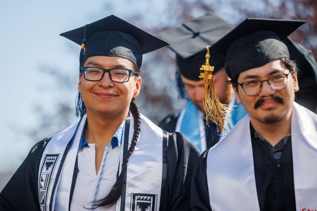 Two graduates wearing white stoles with geometric Indigenous designs smile at the camera. Both wear black caps; one has a gold "2026" tassel. A third graduate is partially visible behind them.
