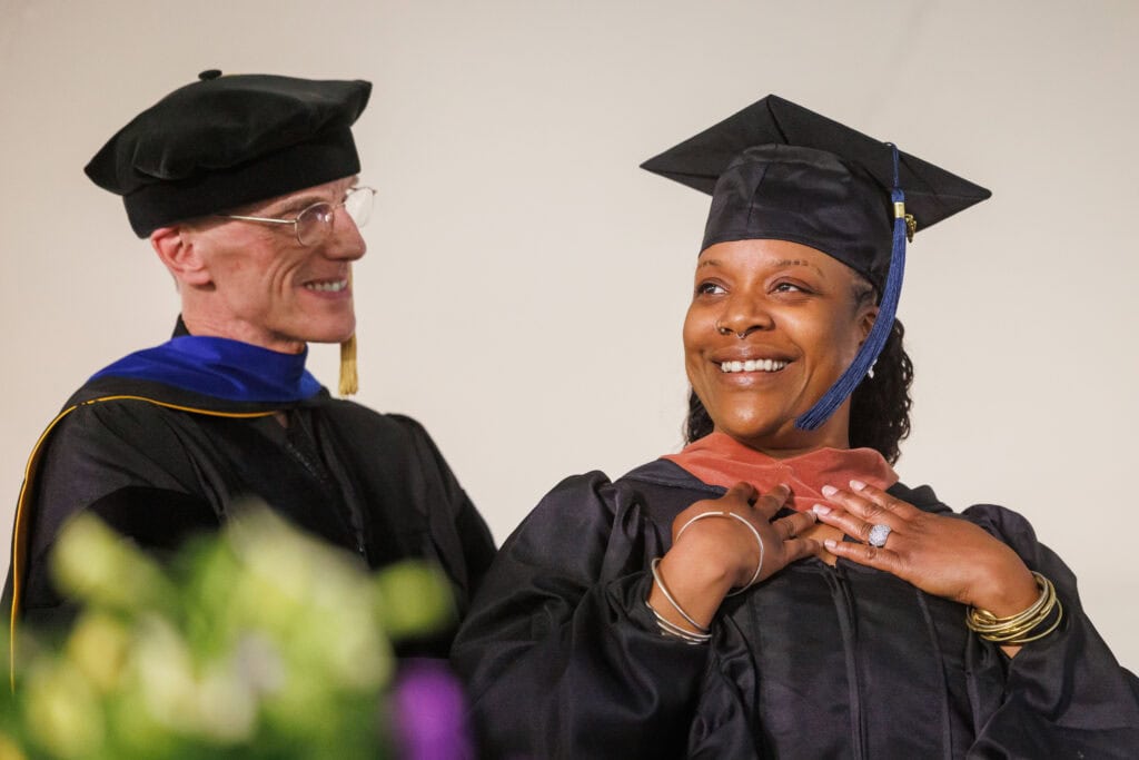 A graduate beams with pride as she adjusts a salmon-colored academic hood draped around her neck during a hooding ceremony. A faculty member in a doctoral tam and blue-and-gold regalia smiles beside her.