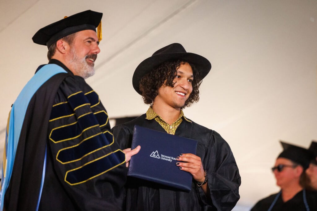 A graduate wearing a wide-brimmed black hat and a patterned shirt beneath his gown smiles proudly while holding a Vermont State University diploma cover. A faculty member in full academic regalia stands beside him.