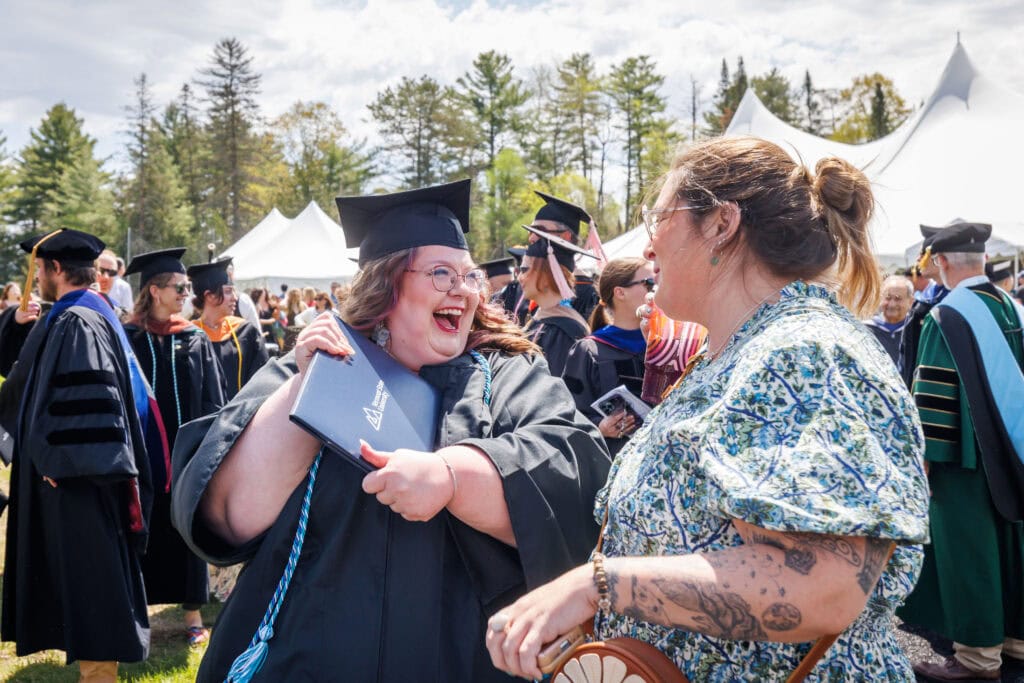 A laughing graduate in cap and gown hugs her diploma cover while sharing a joyful moment with a woman in a floral dress at an outdoor commencement ceremony. White event tents and evergreen trees are visible in the background.