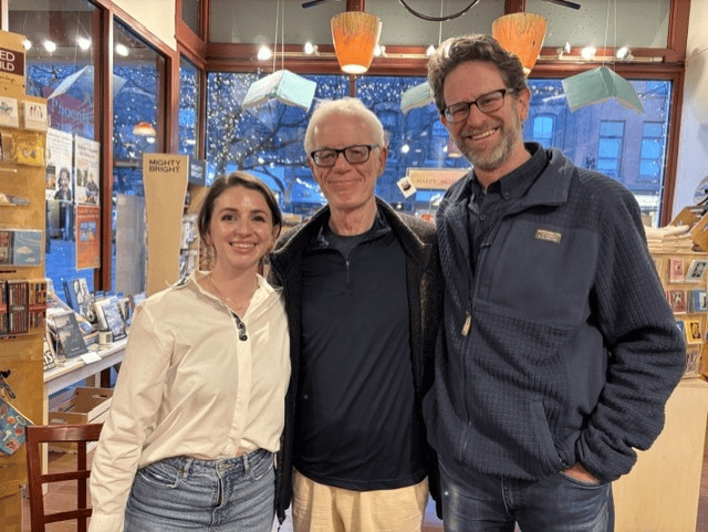Three people smiling and posing together inside Phoenix Books, a bookstore, with shelves of books and a wall of windows visible behind them.