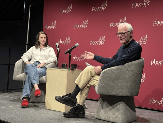 Two people seated in chairs on a stage at Phoenix Books during a speaking event or author interview, with a red branded backdrop behind them.