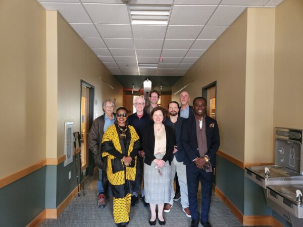 A group of eight people posing together in a university hallway, with a woman in a black-and-yellow print outfit and a woman in a plaid skirt and black blazer standing in the front row, surrounded by six others arranged behind them. A water fountain is visible on the right wall.