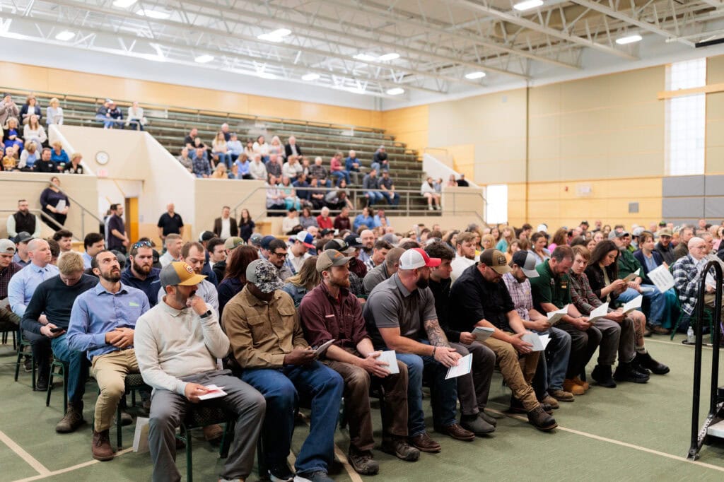 A large group of people sitting in folding chairs, with more seated in bleachers at the back of the room at a graduation ceremony