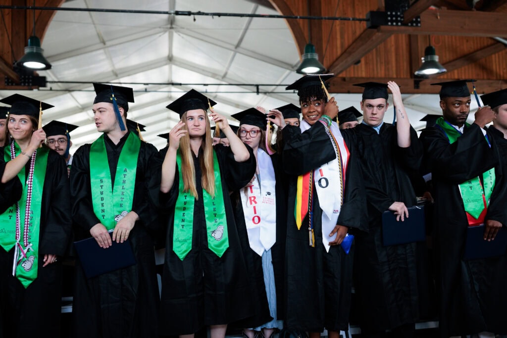 A row of graduates turn their tassels from right to left in unison, marking the official moment of graduation. Many wear identifying stoles, including green "Student Athlete" sashes and white TRIO program stoles, inside a timber-framed venue.