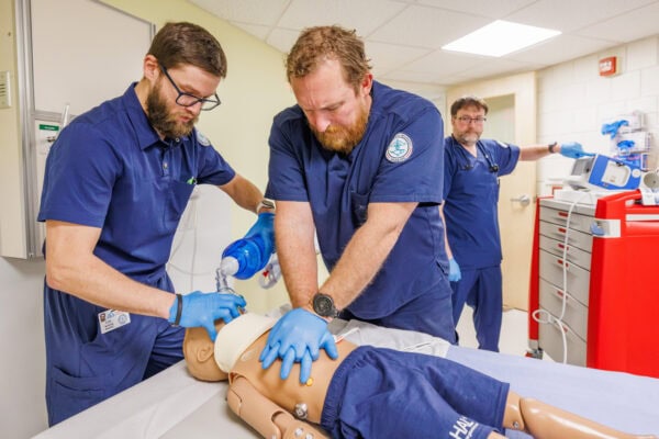 Three nursing students in navy uniforms practice emergency care on a medical training mannequin in a clinical lab. Two students perform chest compressions and manage airflow with a bag-valve mask while a third student operates nearby medical equipment and supplies.