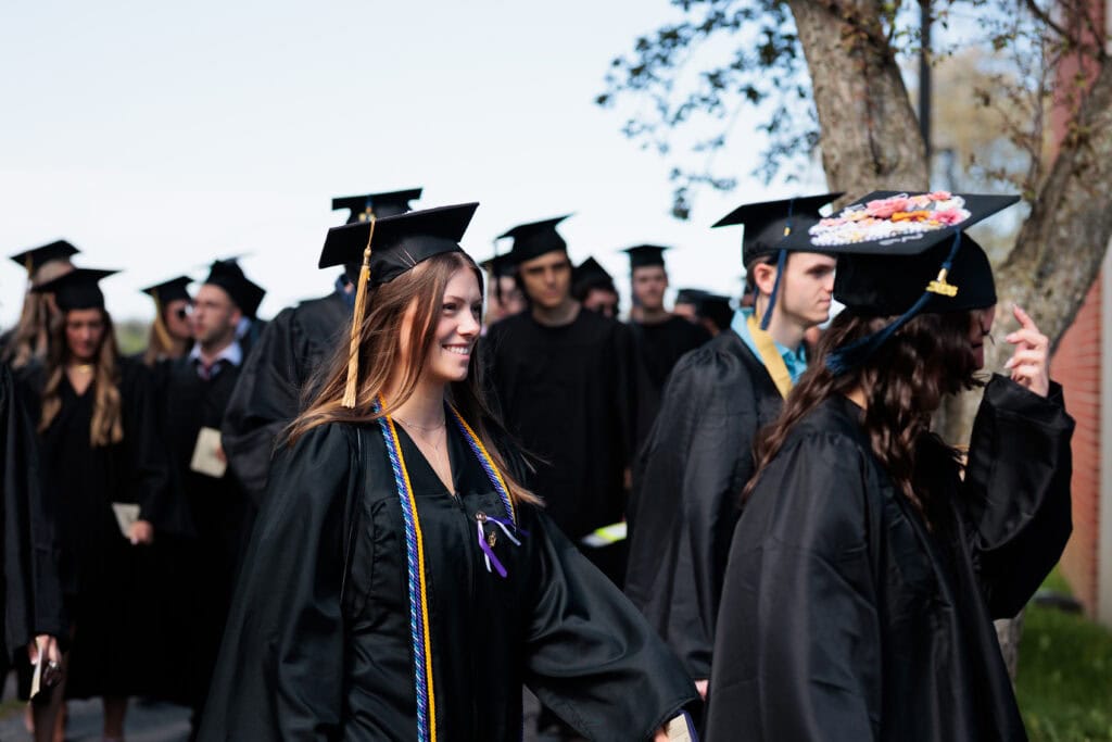 A smiling graduate with long hair and a gold-and-blue honor cord walks in a procession, surrounded by fellow graduates in black caps and gowns. One graduate nearby wears a cap decorated with flowers.